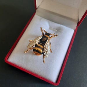 May include: A bee-shaped brooch featuring gold-tone metal, black enamel stripes on the abdomen, and cream-colored enamel wings with gold-tone veins. The insect-shaped pin is displayed inside a small, open jewelry box with a red exterior and white lining.