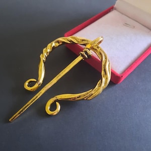 May include: A gold-coloured brooch with a twisted design. The brooch has a curved shape with a long pin. The brooch is displayed in a red box with a white interior, set against a dark grey background.