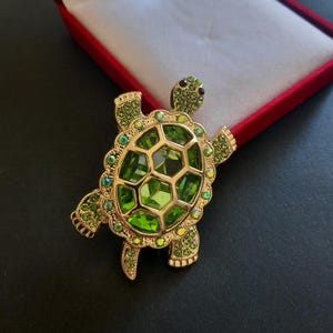 May include: A gold-toned turtle brooch featuring a large, faceted lime green central stone on the shell, surrounded by smaller, sparkling green crystals embedded along the head, legs, and the outer border of the turtle's shell structure.