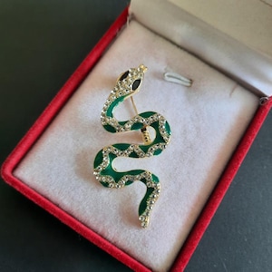 Pode incluir: Broche de cobra dourada com corpo esmaltado verde e detalhes brilhantes de cristal. A cabeça da cobra apresenta olhos de cristal preto. O broche é exibido dentro de uma caixa forrada de veludo vermelho, mostrando seu design.