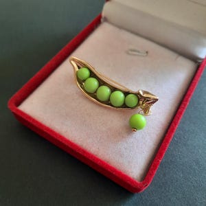 May include: A gold-coloured pea pod brooch with six green peas inside and one dangling below. The brooch is displayed in a red velvet jewellery box with a white interior, set against a dark background.