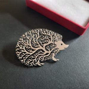 Op de afbeelding: Een zilverkleurige metalen broche in de vorm van een egel. Het lichaam van de egel heeft een ingewikkeld, uitgesneden ontwerp dat doet denken aan vertakkende boomtakken of bladeren, terwijl de kop glad en massief is met een kleine ronde oogopening.