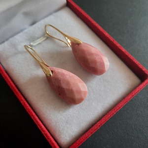 Puede incluir: Un par de pendientes colgantes con piedras rosas talladas en forma de lágrima, suspendidas de ganchos de metal dorado pulido. Los pendientes se presentan dentro de un pequeño joyero cuadrado con exterior rojo y forro interior gris claro.