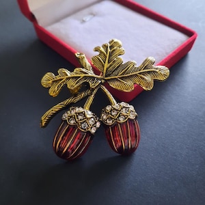 Könnte beinhalten: Goldfarbene Brosche mit einem Eichenblatt und zwei Eicheln. Die Eicheln sind rot mit goldenen Akzenten und mit klaren Kristallen besetzt. Die Brosche liegt in einer roten Samtbox.