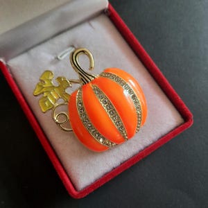 May include: An orange pumpkin brooch with gold accents and clear rhinestones. The brooch has a gold stem and a gold and yellow leaf. The brooch is in a red velvet box with a white interior.