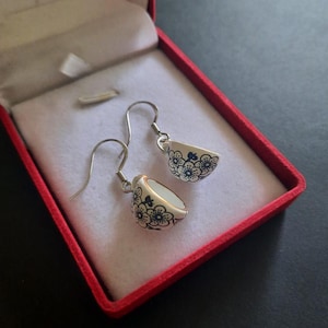 Puede incluir: Un par de pendientes colgantes con forma de cuencos de cerámica blanca en miniatura con un estampado floral azul oscuro. Cada pendiente está sujeto a un gancho de metal plateado. Los pendientes se presentan en un joyero rojo con forro blanco.