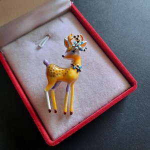 Op de afbeelding: Decoratieve hertenbroche in een rode fluwelen doos. De broche is geel met bruine vlekken, paarse poten en een witte kop. Het heeft een bloemenkrans met blauwe en zwarte accenten. Een uniek accessoire.