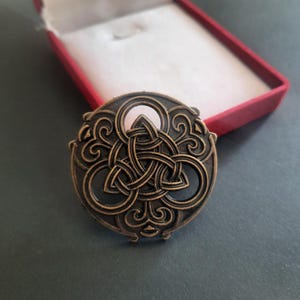 Könnte beinhalten: Eine kreisförmige Brosche aus bronzefarbenem Metall mit einem komplizierten durchbrochenen Design aus einem verschlungenen Triquetra-Knoten, umgeben von geschwungenen Ornamenten. Das Stück hat ein dunkles, antikes Finish.