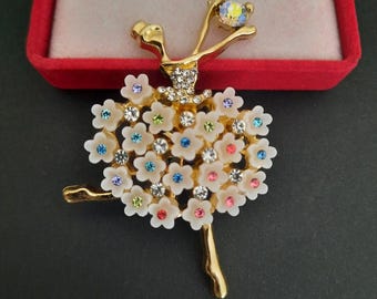 May include: A gold-toned ballerina brooch with a white floral skirt adorned with colorful rhinestones. The ballerina holds a large, iridescent crystal. The brooch is set against a red velvet background.