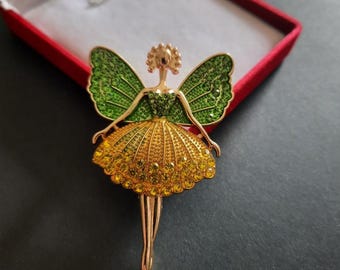 May include: A gold-toned metal brooch shaped like a fairy figure. The fairy has green crystal-encrusted wings, a green bodice, and a flared skirt covered in yellow crystals. The figure has slender legs and a detailed head, all in a polished gold finish.