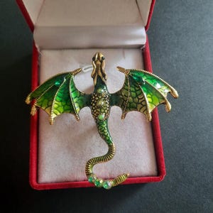 Peut inclure: Broche en forme de dragon avec des ailes et un corps émaillés verts et dorés. Le dragon a une longue queue incurvée et est orné de strass verts. La broche est présentée dans un écrin en velours rouge.