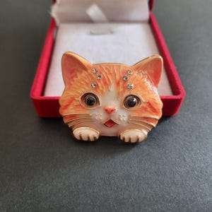 Könnte beinhalten: Eine katzenförmige Brosche mit orangefarbener und weißer Emaille sowie goldfarbenen Metallkonturen. Das Katzengesicht hat große schwarze Augen, eine rosa Nase und vier kleine, klare Strasssteine über den Augen. Zwei kleine Pfoten sind an der Basis sichtbar.