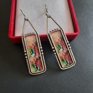 May include: A pair of silver-toned dangle earrings featuring rectangular frames with beaded accents. Each earring contains a central inlay displaying a vibrant, abstract pattern of blended pink, green, and blue colors with a smooth, glossy finish.