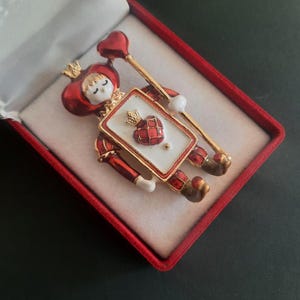 Op de afbeelding: Een goudkleurige metalen broche in de vorm van een speelkaartfiguur met een rode hoed met kroon, die een scepter met een hartje vasthoudt. Het figuur heeft een wit gezicht en een rechthoekig lichaam met een rood hartsymbool op een witte achtergrond.