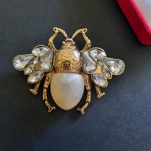 Könnte beinhalten: Goldfarbene Bienenbrosche mit klaren Kristallakzenten auf den Flügeln und einem perlmuttartigen Körper. Die Brosche hat aufwendige Details und eine strukturierte Oberfläche, die einen luxuriösen Look erzeugt. Dieses Schmuckstück eignet sich, um jedem Outfit einen Hauch von Eleganz zu verleihen.