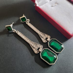 Puede incluir: Un par de pendientes largos con montura de metal plateado engastada con pedrería blanca transparente. Cada pendiente tiene una pequeña piedra verde cuadrada en el poste y una piedra verde rectangular más grande en la base.