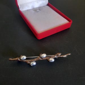 May include: A silver-coloured brooch shaped like a branch, adorned with five pearl-like beads. The brooch is set against a black surface, with a red jewellery box in the background.