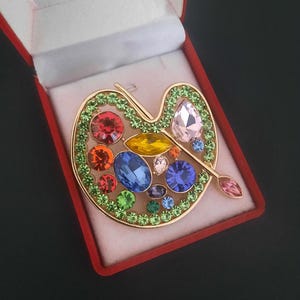 Puede incluir: Broche dorado con forma de paleta de artista, incrustado con gemas coloridas. La paleta está bordeada con cristales verdes y presenta un pincel. El broche se presenta en una caja de terciopelo rojo.