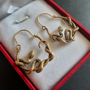 Puede incluir: Un par de pendientes de aro en tono dorado con un diseño de serpiente texturizada. Cada pendiente tiene forma de serpiente enroscada con un patrón de escamas detallado, formando una estructura de aro circular que se cierra con un alambre fino.