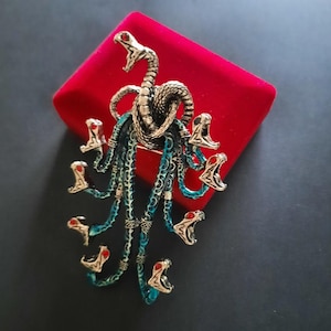 Puede incluir: Broche detallado con un diseño de serpiente de múltiples cabezas. El broche tiene un acabado plateado y dorado con detalles turquesa y rojo. Las cabezas de serpiente tienen la boca abierta y el broche está sobre un fondo de terciopelo rojo.
