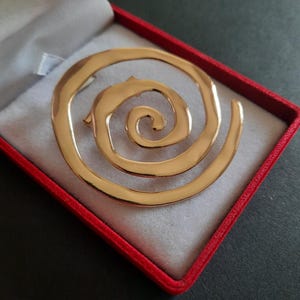 May include: A gold-toned metal brooch featuring a continuous spiral design with a smooth, polished surface. The circular ornament has a tapering end that curves inward toward the center, creating a fluid, open-work shape with a reflective finish.