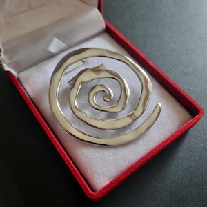 Peut inclure: Une broche en métal argenté présentant un motif en spirale continue avec une surface lisse et polie. La broche est présentée à l'intérieur d'une boîte à bijoux rectangulaire dotée d'un extérieur rouge et d'une doublure intérieure claire.