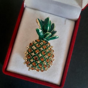 Puede incluir: Broche de piña dorada con parte superior de esmalte verde y detalles de cristal color ámbar. La broche está colocada en un joyero de terciopelo rojo.