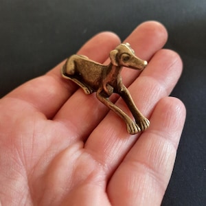 Könnte beinhalten: Eine kleine, antik wirkende Metallfigur in Bronzetönen, die einen Hund in liegender Ruheposition darstellt. Die Hundeform zeichnet sich durch einen schlanken Körper, spitze Ohren und ausgestreckte Vorderpfoten mit einer texturierten Oberfläche aus.