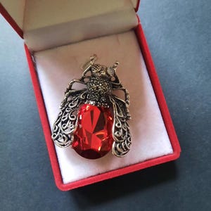 Puede incluir: Broche plateado con forma de escarabajo, con una gran gema roja facetada como cuerpo. Las alas están detalladas con filigrana calada. El broche se presenta en una caja forrada de terciopelo rojo.