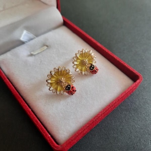 Op de afbeelding: Een paar oorknopjes in de vorm van een gele margriet met witte bloemblaadjes en een klein rood lieveheersbeestje met zwarte stippen in het midden. De oorbellen zijn gemaakt van goudkleurig metaal en liggen in een rood fluwelen doosje.