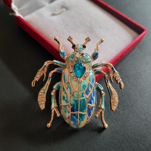 Op de afbeelding: Een kevervormige broche met een goudkleurig metalen frame en blauwe, turquoise en witte emaille details. Het insectenlichaam is versierd met een centrale druppelvormige blauwe kristal, kleinere heldere strass-steentjes en filigraanpatronen.