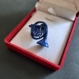 Op de afbeelding: Een glanzende, diepblauwe emaille reversspeld in de vorm van een hoorn. De speld in de vorm van een muziekinstrument heeft gedetailleerde opgerolde buizen en een uitlopende beker, gepresenteerd in een rood juwelendoosje met witte voering.