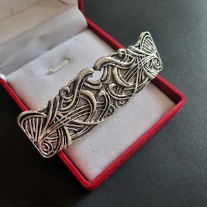 Puede incluir: Un pasador de pelo rectangular de metal plateado con un intrincado patrón en relieve de líneas entrelazadas y motivos en forma de abanico. El accesorio se presenta dentro de un pequeño joyero rojo abierto con un interior de color blanco.