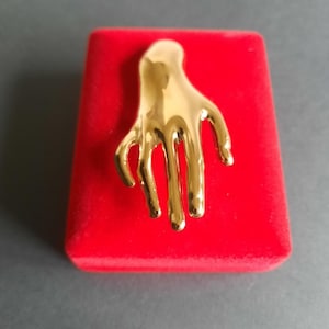 Pode incluir: Um broche de tom dourado em forma de mão estilizada e alongada, com superfícies lisas e reflexivas. A peça apresenta cinco dedos finos e afilados apontando para baixo, criando uma forma metálica fluida sobre uma base de veludo vermelho.