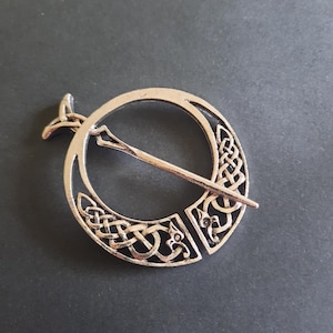 Könnte beinhalten: Silberfarbene keltische Knotenbrosche. Die runde Brosche weist komplizierte Knotenmuster und einen Nadelverschluss auf. Das Design umfasst einen zentralen Ring mit einem dekorativen Muster und eine separate Nadel zur Befestigung der Brosche.