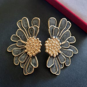 Könnte beinhalten: Ein Paar Statement-Ohrringe mit floralem Design. Jeder Ohrring hat eine goldfarbene Mitte mit kleinen, runden Akzenten, umgeben von dunkelgrauen Blütenblättern mit goldfarbenen Umrissen. Die Ohrringe sind vor einem schwarzen Hintergrund platziert.