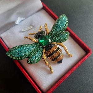 May include: A bee-shaped brooch featuring gold-tone metal legs and antennae, a large emerald-green central crystal, wings encrusted with small teal and green rhinestones, and an abdomen striped with black enamel and amber-colored crystals.