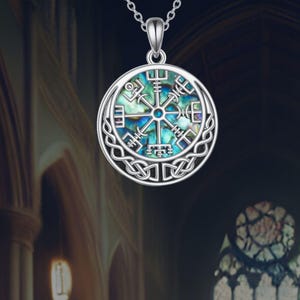 Puede incluir: Un collar con colgante plateado con un colgante circular que muestra un diseño de brújula vikinga. El colgante tiene un centro iridiscente azul y verde y está enmarcado por un borde de nudo celta. El collar cuelga de una fina cadena.