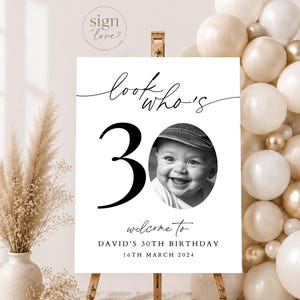 May include: A white rectangular birthday sign on a wooden easel featuring the text "look who's 30" in black script, a large black number 30, a circular black and white photo of a smiling baby, and the text "welcome to DAVID'S 30TH BIRTHDAY 16TH MARCH 2024".