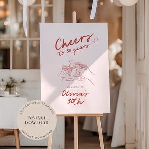May include: A white sign on a wooden easel with the text "Cheers to 30 years" in red script. Below the text is an illustration of three hands toasting with glasses. The sign reads "Welcome to Olivia's 30th".