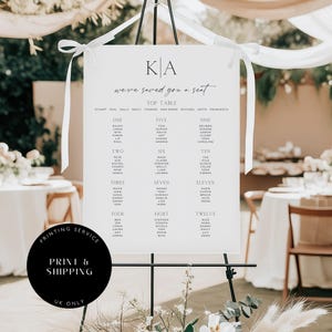 May include: A white wedding seating plan with black text, displayed on a black easel. The chart features the initials "KA" at the top, followed by the words "we've saved you a seat" and guest names organised by table number. White ribbons secure the chart.