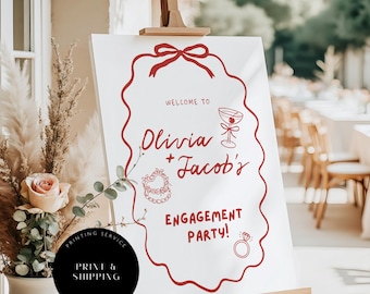 May include: A white sign on a wooden easel with red text and illustrations. The sign reads "WELCOME TO Olivia + Jacob's ENGAGEMENT PARTY!" with a bow, a cocktail, a heart-shaped wreath, and a ring illustration. A black circle with text is in the lower left.