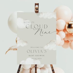 May include: A baby shower welcome sign on an easel. The sign is light grey with the words "We're on Cloud Nine" and "Welcome to Olivia's Baby Shower 03.04.2025". The sign is decorated with cloud illustrations. Balloons in white, gold, and peach surround the sign.