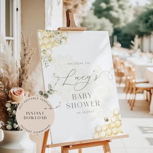 Könnte beinhalten: Ein weißes Willkommensschild für eine Babyparty auf einer Holzstaffelei. Das Schild zeigt ein Wabenmuster mit floralen Akzenten und die Worte "Willkommen zu Lucys Babyparty" in eleganter Schrift. Eine kleine Bienen-Grafik ist ebenfalls enthalten.