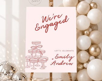 May include: A rectangular sign on a wooden easel with a light pink background and red text reading We're Engaged, Let's Celebrate, Emily + Andrew. A red line drawing of a stacked champagne coupe tower is featured on the left side of the sign.