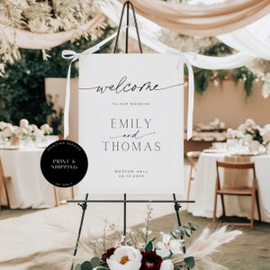 May include: A white wedding sign on a black easel, with the words "welcome" and the names "Emily and Thomas" in elegant script. The sign also includes the wedding date and venue.