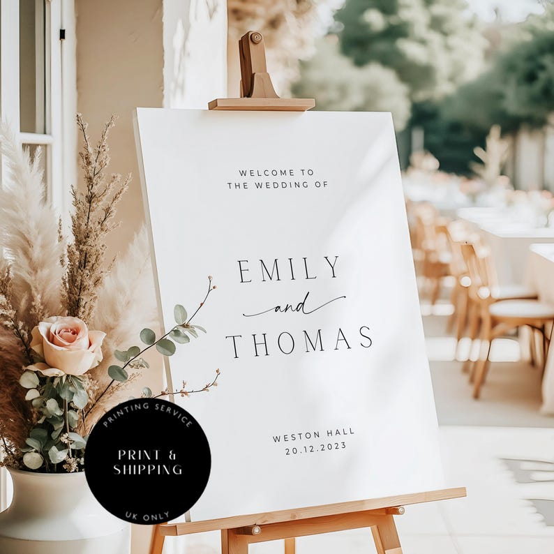 May include: A white wedding welcome sign on a wooden easel. The sign reads "Welcome to the Wedding of Emily and Thomas" with the date 20.12.2023 and location Weston Hall. A black circle with text "PRINT & SHIPPING UK ONLY" is in the lower left.