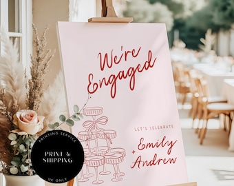 May include: A light pink sign on a wooden easel with the words "We're Engaged" in red script. Below, a graphic of champagne glasses and the names "Emily + Andrew". A black circle reads "Printing Service" and "UK Only".