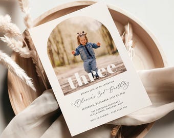 Op de afbeelding: Een witte uitnodigingskaart met een foto van een kind in een blauw spijkerjack en spijkerbroek. De kaart heeft een witte boog met het woord "three" in vetgedrukte witte letters. De tekst "Please join us to celebrate Olivia's 3rd Birthday" is geschreven in cursief schrift. De uitnodiging bevat de datum, tijd, locatie en RSVP-informatie.