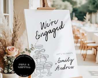 May include: A white sign on a wooden easel with the words "We're Engaged" in black script. Below, it reads "Let's Celebrate" and the names "Emily + Andrew". A black circle says "PRINTING SERVICE" and "UK ONLY".